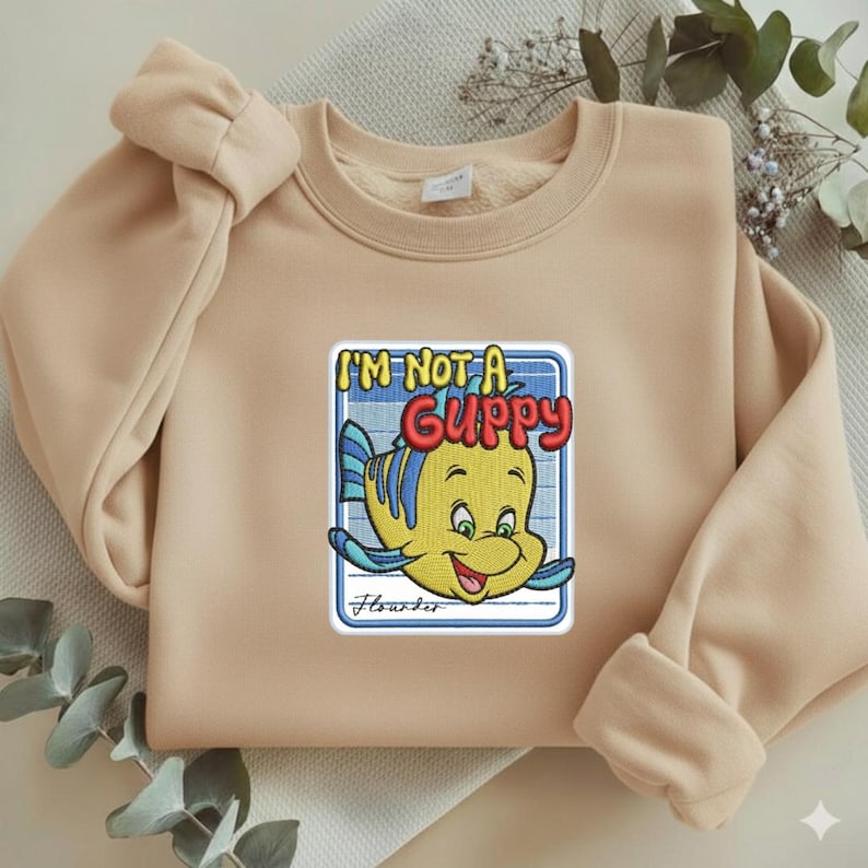 May include: A beige crewneck sweatshirt featuring a central embroidered patch of a yellow and blue fish character. The text reads I'M NOT A GUPPY in bold red letters above the fish, with the name Flounder written in cursive script below.