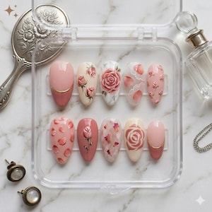 May include: A set of ten artificial nails in a clear plastic case. The nails are painted with various floral designs, including roses and petals, in shades of pink, white, and gold. Some nails feature 3D rose accents. The nails are oval-shaped.