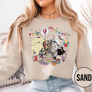 May include: Sand-colored sweatshirt featuring a colorful graphic of characters from The Nightmare Before Christmas celebrating a birthday. The design includes a cake, balloons, and the number 4. The word "SAND" is printed in a circle on the bottom right.