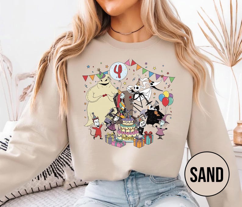 May include: Sand-colored sweatshirt featuring a colorful graphic of characters from The Nightmare Before Christmas celebrating a birthday. The design includes a cake, balloons, and the number 4. The word "SAND" is printed in a circle on the bottom right.