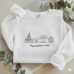 May include: A white long-sleeve crewneck sweatshirt featuring a black line art graphic of four iconic theme park landmarks and the text "Happiest place on earth" printed in a cursive script font centred on the chest area of the garment.