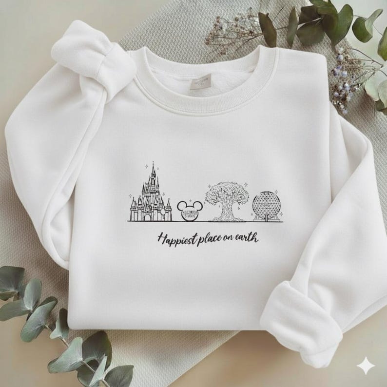 May include: A white long-sleeve crewneck sweatshirt featuring a black line art graphic of four iconic theme park landmarks and the text "Happiest place on earth" printed in a cursive script font centred on the chest area of the garment.