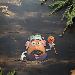 May include: A Halloween-themed Mr. Potato Head figure, dressed as Frankenstein, holding a jack-o'-lantern on a stick. The figure has a green headpiece, a black mustache, and a purple vest. The background is a dark wooden surface.