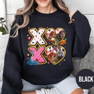 May include: Black sweatshirt featuring a graphic design with the letters "XO" in gold glitter and pink. The design includes cartoon characters in heart-shaped frames. The word "BLACK" is printed on a black circle.