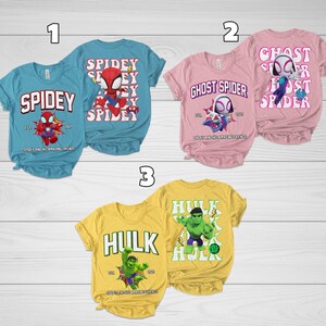 Puede incluir: Una colecci&oacute;n de tres camisetas de manga corta con dise&ntilde;os de superh&eacute;roes. Las camisetas son de color azul claro, rosa y amarillo. Cada camiseta tiene un gr&aacute;fico y texto de superh&eacute;roe diferente, incluyendo "Spidey", "Ghost Spider" y "Hulk".