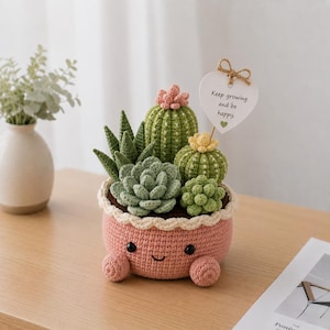 May include: A crocheted succulent garden in a pink pot with a smiling face and small feet. The garden features various green and light green cacti and succulents, with a heart-shaped tag that reads "Keep growing and be happy."