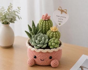 Puede incluir: Un jardín de suculentas de ganchillo en una maceta rosa con una cara sonriente y pies pequeños. El jardín presenta varios cactus y suculentas verdes y verde claro, con una etiqueta en forma de corazón que dice "Keep growing and be happy."
