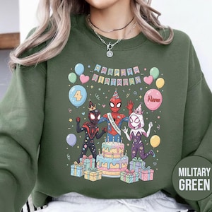 May include: Military green sweatshirt featuring a colorful graphic of Spider-Man characters celebrating a birthday. The design includes a cake, balloons, and the text "Happy Birthday". The number "4" is also displayed.