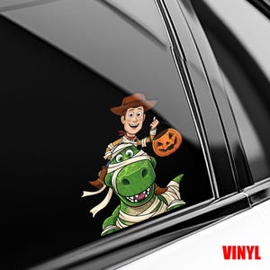 May include: A vinyl decal featuring Woody from Toy Story riding a green dinosaur wrapped in bandages. Woody holds a jack-o'-lantern. The word "VINYL" is printed in red.