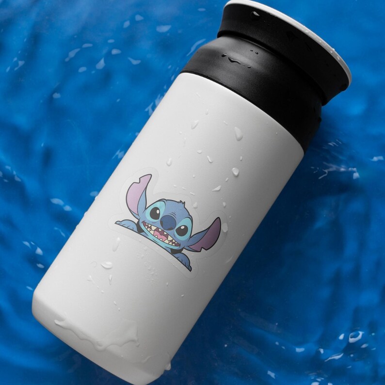 May include: White insulated tumbler with a black lid featuring a cartoon image of Stitch. The blue and pink character is smiling. The tumbler is cylindrical and appears to be made of stainless steel.