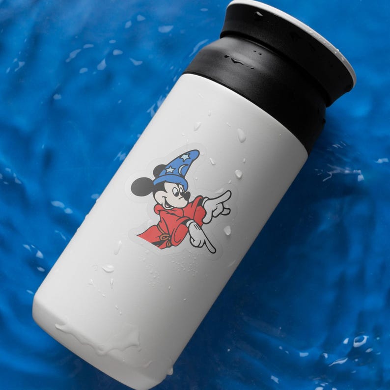 May include: White insulated tumbler with a black lid, featuring a colorful sticker of Mickey Mouse in a wizard outfit. The cartoon character is wearing a blue hat with stars and a red robe, pointing with his finger.