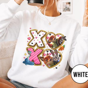 May include: White sweatshirt featuring a design with the letters "XO" in pink and white, surrounded by gold glitter. The design includes cartoon characters in heart-shaped frames, with small hearts scattered around the letters.