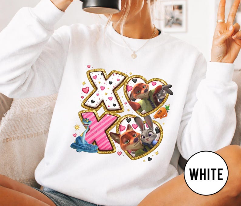 May include: White sweatshirt featuring a design with the letters "XO" in pink and white, surrounded by gold glitter. The design includes cartoon characters in heart-shaped frames, with small hearts scattered around the letters.