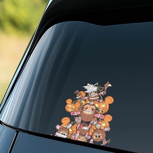 May include: A colorful car decal featuring a cartoon illustration of several characters with pumpkin heads and wrapped in bandages. The characters are holding weapons and are stacked on top of each other. The decal is on a car window.