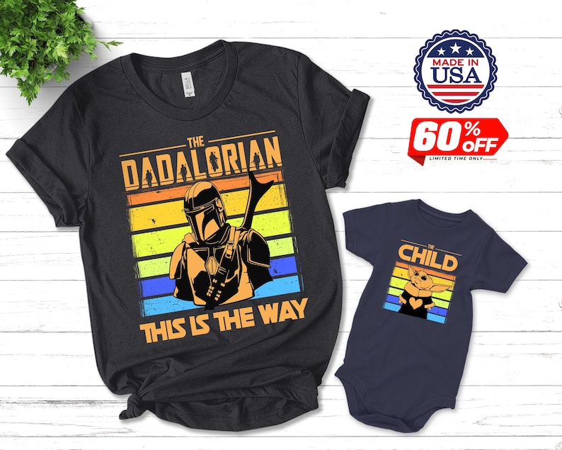 May include: Black t-shirt with "THE DADALORIAN" and "THIS IS THE WAY" text, featuring a Mandalorian graphic. A navy blue onesie with "CHILD" text and a Grogu graphic is also shown. Made in USA.