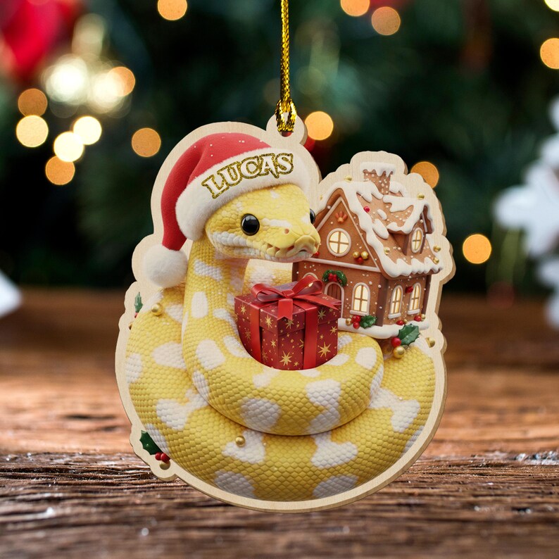 May include: A wooden Christmas decoration depicting a yellow snake with white spots wearing a Santa hat with the name "LUCAS". The snake is coiled around a red gift box and a gingerbread house. The ornament hangs from a gold string.