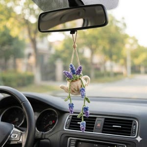 May include: A crocheted car charm featuring a small teapot filled with purple and blue lavender flowers. The teapot is beige with green accents and hangs from a wooden bead and string, suspended from the rearview mirror.