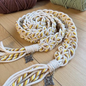 Op de afbeelding: Een handgemaakte macramé riem in crème, geel en bruin. De riem heeft een gevlochten ontwerp met decoratieve kwastjes en zilveren Keltische knoopbedels. De riem is gemaakt van dik, duurzaam touw.