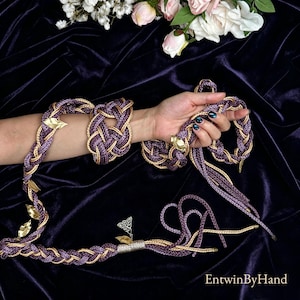 Op de afbeelding: Een verzameling gevlochten accessoires in paars en goud, waaronder een armband, een riem en een hoofdband. De hoofdband heeft gouden bladeren. De accessoires worden getoond op een donkerpaarse fluwelen ondergrond.