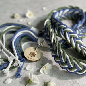 Op de afbeelding: Gevlochten touwlijn in blauw, groen en wit. De lijn heeft een houten label met een sneeuwvlokontwerp en een metalen sluiting. Witte bloemen zijn verspreid.