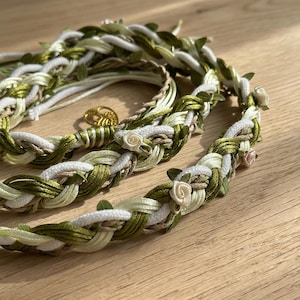 Op de afbeelding: Gevlochten hoofddeksel met witte, groene en beige strengen, versierd met kleine witte en roze stoffen rozen en groene bladeren. Een goudkleurige bedel is ook zichtbaar. Geschikt voor bruiloften of speciale gelegenheden.