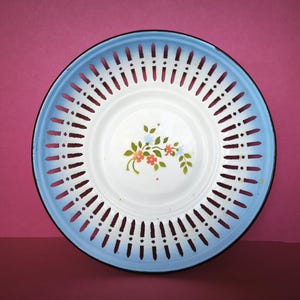 Op de afbeelding: Een witte emaille kom met een blauwe rand en een bloemenmotief. De kom heeft een decoratief opengewerkt patroon rond de rand.