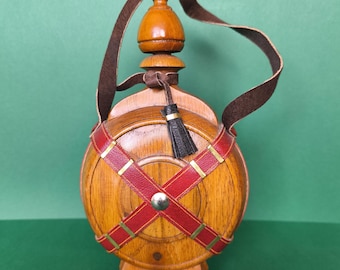 Könnte beinhalten: Eine Holzflasche mit einem braunen Lederriemen und einem dekorativen Holzverschluss. Die Flasche ist von roten Lederriemen mit goldenen Akzenten umgeben. Eine kleine schwarze Quaste hängt am Verschluss. Die Flasche steht auf einem Holzsockel.