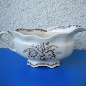 May include: A white ceramic gravy boat featuring a floral pattern in grey and black. It has a handle and a spout, and rests on a decorative base. The rim is trimmed with a grey and black pattern.