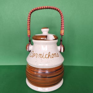 May include: A white ceramic jar with a brown and white striped design. The jar has a brown wicker handle and says "Cornichons" in brown lettering.
