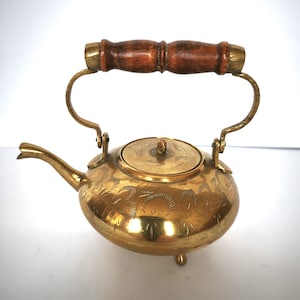 Puede incluir: Pequeña tetera de latón vintage con asa de madera. La tetera tiene un cuerpo redondo, un pico y una tapa. El asa es de madera oscura y está unida a la olla con soportes de latón. El cuerpo de la tetera está decorado con patrones florales grabados.