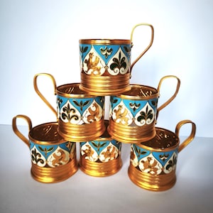 Pode incluir: Conjunto de seis copos de shot de metal dourado com desenhos de esmalte azul, branco e verde. Cada copo apresenta um animal estilizado e motivos florais.