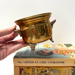 Puede incluir: Un pequeño recipiente de latón pulido con cuerpo redondeado, borde ancho y dos asas laterales. El recipiente cuenta con tres patas decorativas y un acabado dorado metálico. Se sitúa sobre dos libros apilados, uno titulado The American Girl Cookbook.
