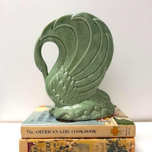 May include: A sage green ceramic swan figurine with a glossy finish, featuring stylised wing feathers and a curved neck, resting on top of two stacked vintage books titled The American Girl Cookbook and The Side of Innocence.