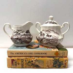 May include: A white ceramic creamer and a lidded sugar bowl with brown transferware landscape patterns featuring houses, trees, and hills. The pieces have curved handles and are displayed on top of two stacked vintage hardcover books.