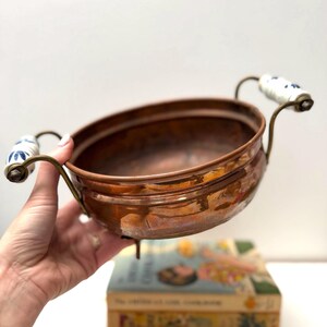 Peut inclure: Un bol rond en cuivre avec une finition métallique polie, doté de deux poignées en laiton courbées fixées à des prises en céramique blanche ornées de motifs floraux bleus. Le bol présente une large ouverture et une base arrondie.