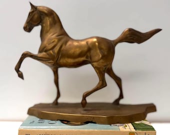 Puede incluir: Una figura de caballo color bronce en pose de trote, montada sobre una base plana e irregular. El caballo está colocado sobre dos libros apilados, uno titulado The American Girl Cookbook y el otro Third Side of Innocence.