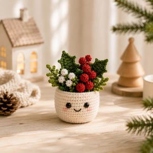 May include: A small, cream-colored crocheted pot featuring a stitched smiling face with black eyes and pink cheeks. The pot contains a festive arrangement of green crocheted holly leaves, white berries, and clusters of bright red berries.
