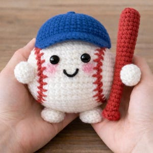 May include: A handmade crocheted baseball character with a smiling face, wearing a blue cap, and holding a red bat. The baseball has red stitching and rosy cheeks. A charming, handcrafted item.
