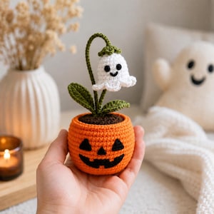 Puede incluir: Una pequeña planta de ganchillo con una flor blanca en forma de fantasma con ojos y sonrisa negros, unida a un tallo verde con dos hojas. La planta está en una maceta redonda de ganchillo naranja con forma de calabaza y cara negra bordada.