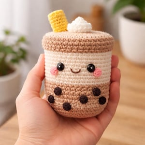Op de afbeelding: Een gehaakte boba thee knuffel in beige en bruin, met een lachend gezicht, roze wangen en zwarte kraalogen. Het heeft een gele rietje en een witte slagroom topping. De bodem heeft donkerbruine bobas.