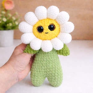 Könnte beinhalten: Handgemachte Häkel-Gänseblümchen-Puppe mit weißem Blütenkopf, gelber Mitte und grünem Stiel und Körper. Die Puppe hat große schwarze Augen und ein lächelndes Gesicht. Die Puppe ist ein skurriles, weiches Spielzeug.