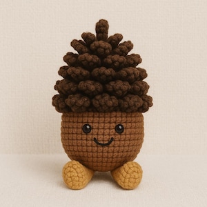 May include: A crocheted acorn plush toy with a smiling face. The top is dark brown, resembling a pinecone, and the body is light brown. Two small, yellow-brown feet are at the bottom. The toy is handmade.