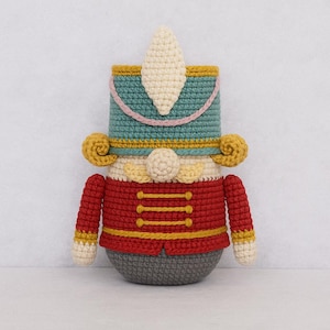 Peut inclure: Poupée casse-noisette au crochet faite à la main. La poupée présente une veste rouge avec des boutons et des bordures dorées, un chapeau bleu sarcelle avec un panache blanc et une base grise. La poupée a des accents jaunes et des mains et des pieds crème.