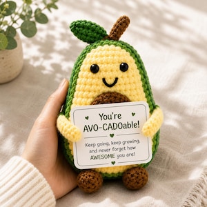 Puede incluir: Un peluche de aguacate tejido a ganchillo con una cara sonriente, piel verde y un hueso marrón. Sostiene un letrero con el texto "You're AVO-CADOable!" y un mensaje inspirador. El peluche tiene pequeños pies marrones y una hoja verde.