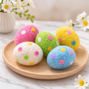 Op de afbeelding: Een houten bord toont zes kleurrijke, gespikkelde paaseieren. De eieren zijn in tinten roze, groen, geel, wit en blauw, elk versierd met contrasterende stippen. De eieren zijn gemaakt van een zacht materiaal.