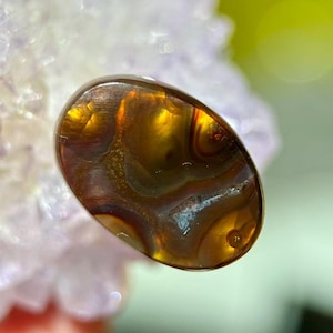 Op de afbeelding: Een gepolijste, ovale edelsteen met een warme kleurenpalet. De steen heeft wervelende patronen van amber, bruin en goud, met doorschijnende gebieden. De edelsteen is waarschijnlijk een decoratief item of een onderdeel van sieraden.