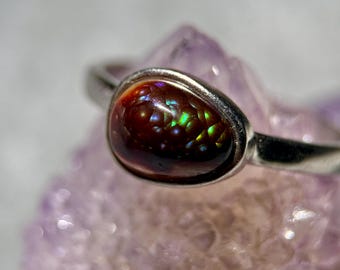 Pode incluir: Um anel de prata com uma pedra preciosa de ágata de fogo lisa e arredondada. A pedra apresenta uma cor base castanho-avermelhada profunda com reflexos iridescentes de luz verde e dourada que brilham na sua superfície polida e abobadada.