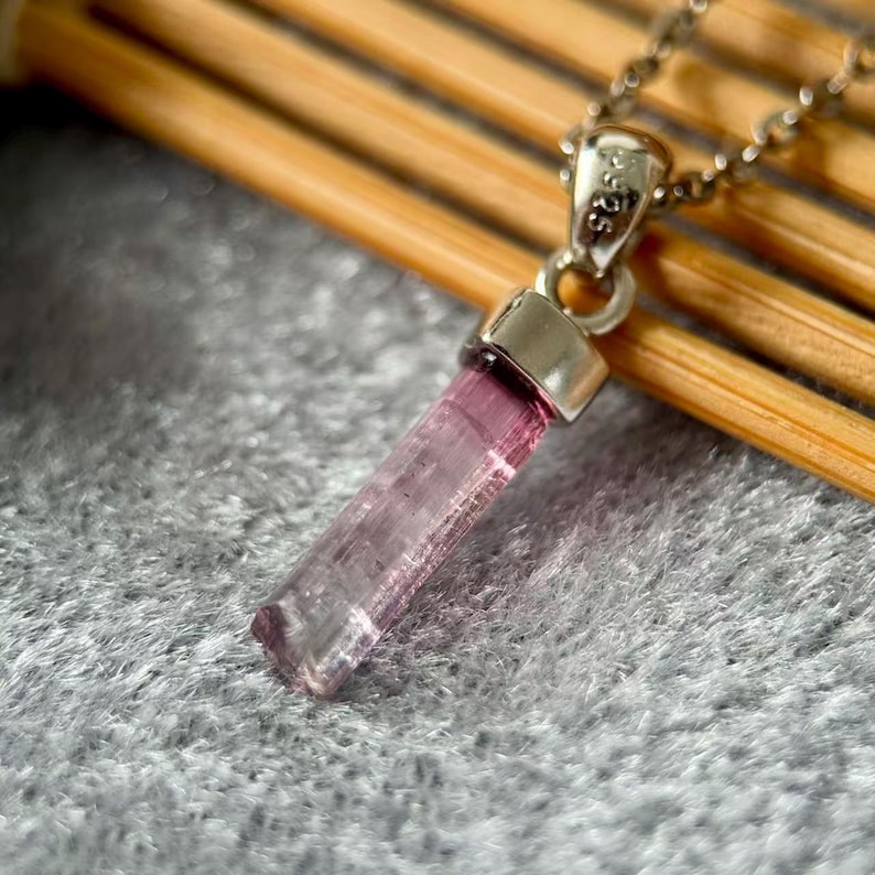 May include: A pendant necklace featuring a raw, elongated pink tourmaline crystal suspended from a silver bail stamped with 925. The crystal has a translucent, faceted appearance with vertical striations, attached to a delicate silver chain.