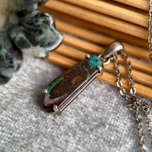 May include: A silver necklace featuring a rectangular pendant with a dark brown opal. The opal has iridescent blue, green, and orange flashes. The pendant is encased in a silver setting and hangs from a delicate silver chain.