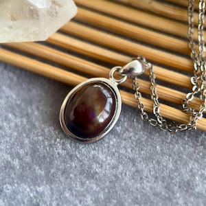 Op de afbeelding: Zilveren ketting met een ovale hanger met een donkere, iriserende edelsteen. De hanger is gezet in een zilveren bezel en hangt aan een fijne ketting. De edelsteen toont bruine, paarse en gouden kleuren.